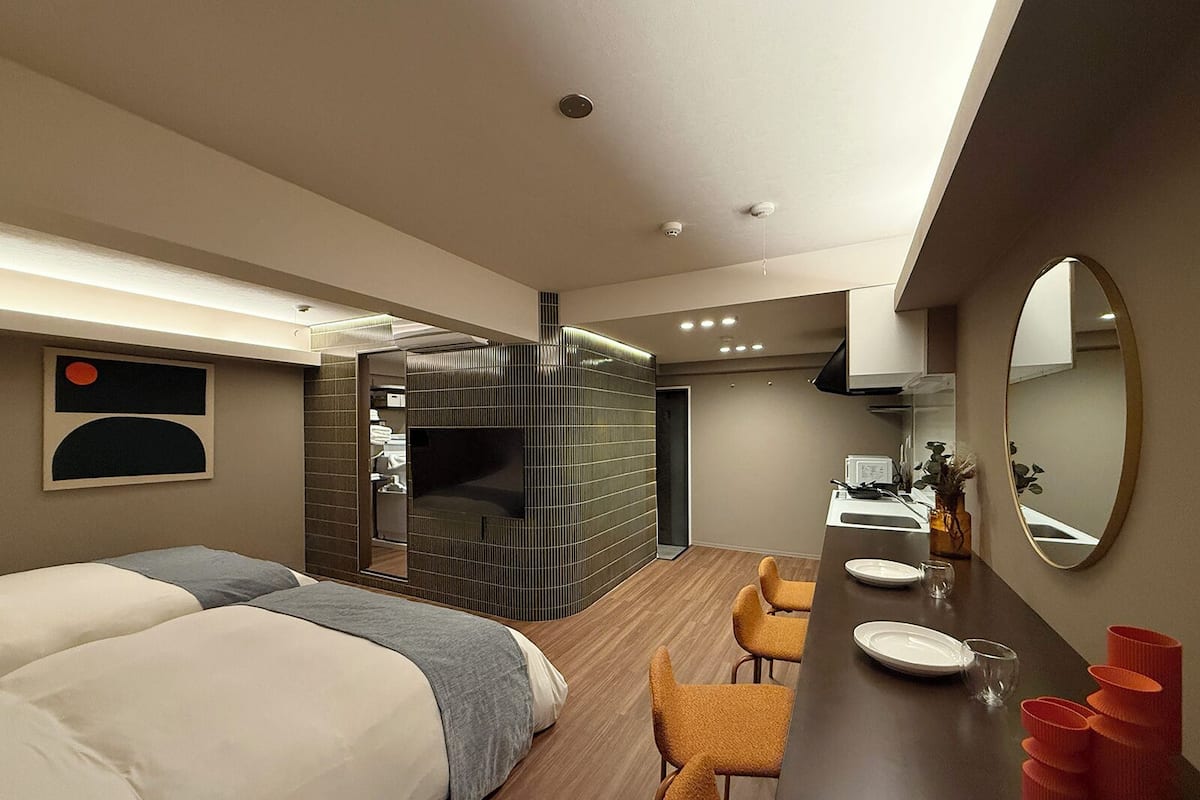 A modern hotel room with a large bed, a small bathroom, a dining area with a table and chairs, and a large mirror.