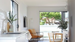 Una cocina moderna con armarios blancos, un comedor con sillas de madera, un ventanal con vista a árboles y una lámpara colgante.