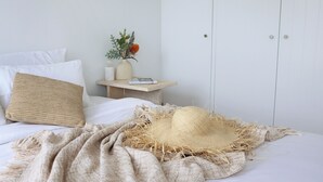 Una cama bien hecha con un sombrero de paja, un cojín tejido y una mesita de noche con un libro y un jarrón de flores.