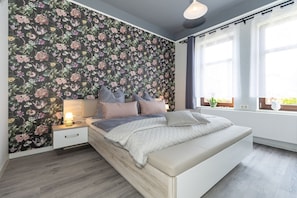 A bedroom with a floral wallpaper, a bed with a white headboard, a bedside table with a lamp, and a window with curtains. - Blumentraum Vacation Rental with 2 Bathrooms - Bright "Blumentraum" Vacation Rental in Kühlungsborn (Kühlungsborn)