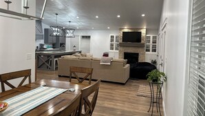 A modern kitchen with a dining area, a living room with a sofa and a fireplace, and a TV mounted above the fireplace.