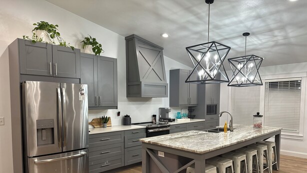 A modern kitchen with grey cabinets, a stainless steel refrigerator, a marble island with bar stools, and pendant lights.