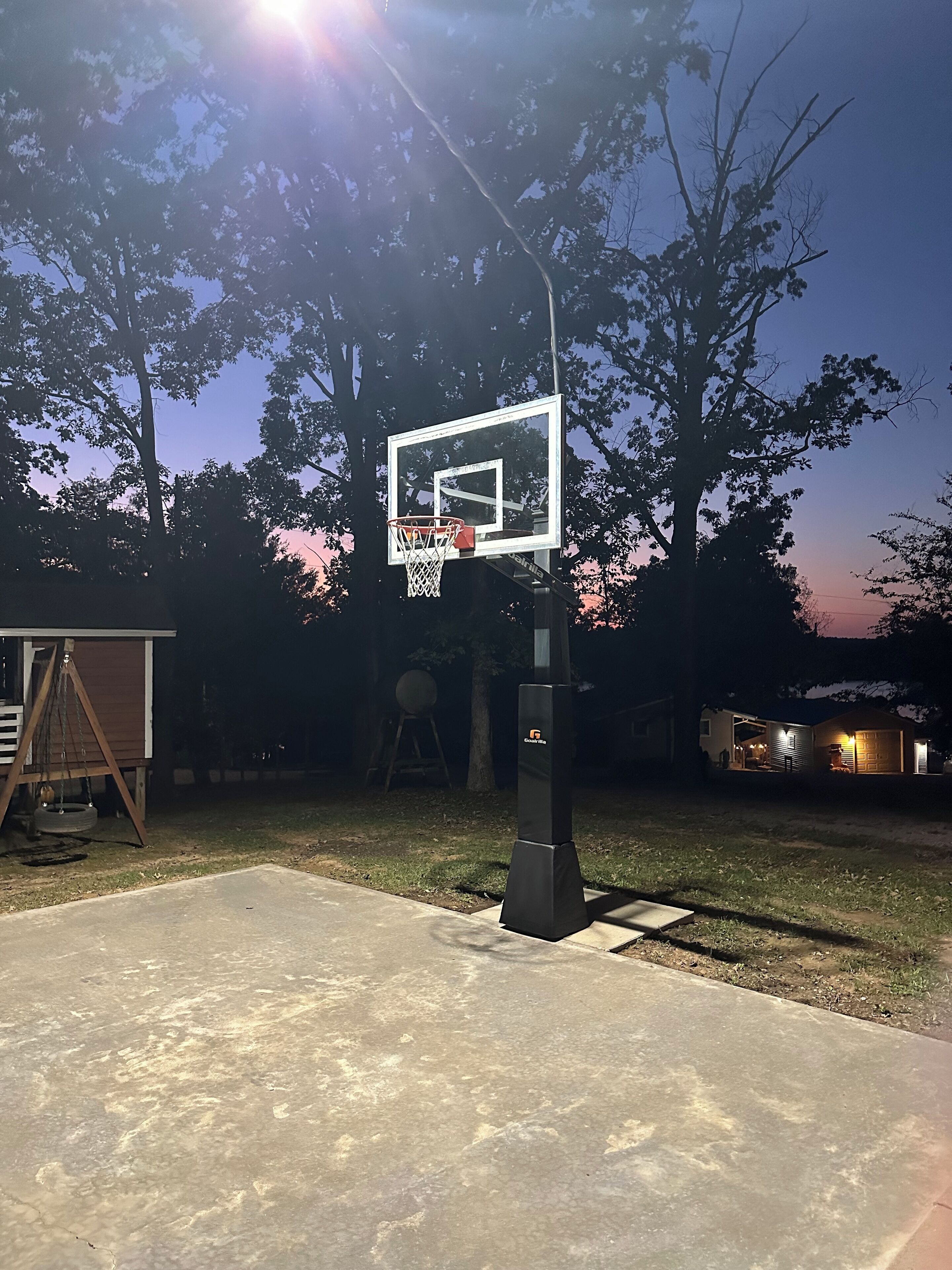 En basketkorg utomhus, placerad på en bakgård med en gunga och ett hus i bakgrunden.