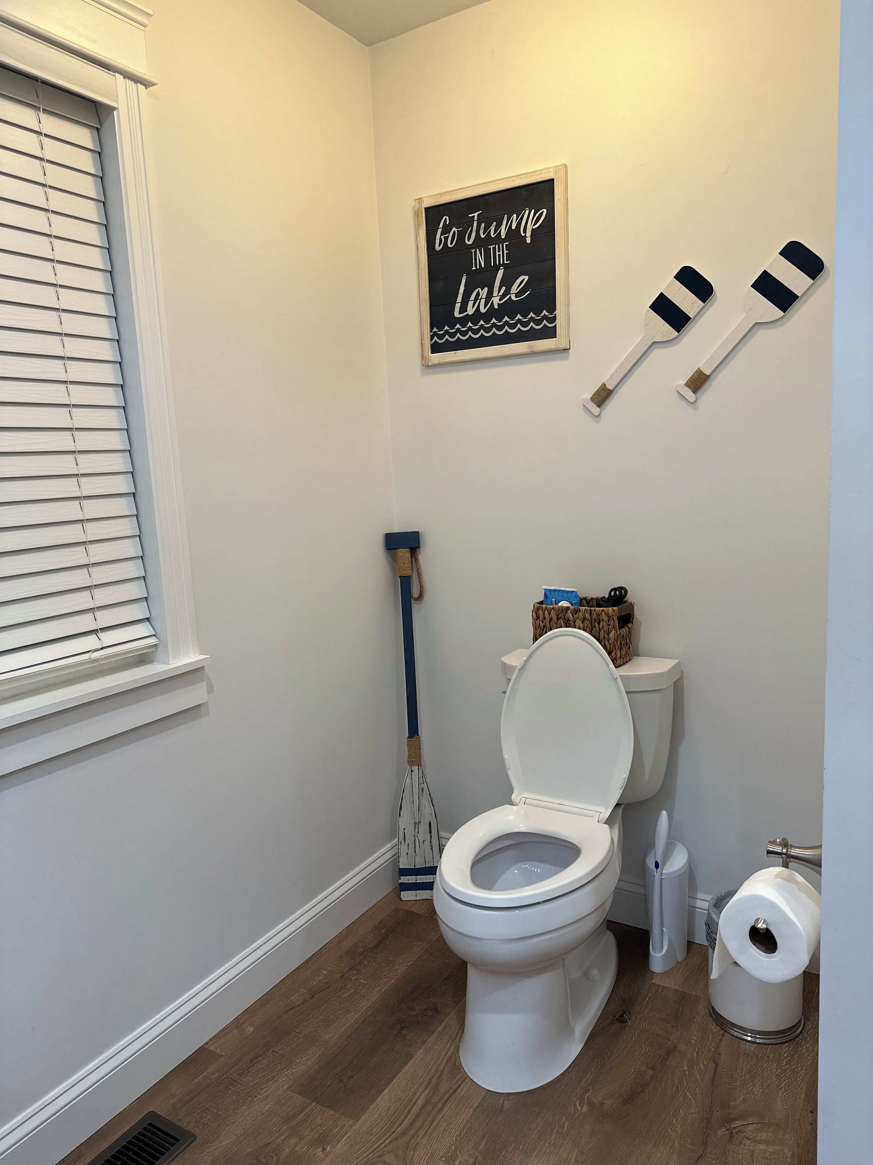 Un baño con inodoro, suelo de madera, ventana con persianas, una escoba, un letrero que dice "Lánzate al Lago" y dos remos de madera.