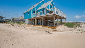 Una casa frente a la playa con balcón, rodeada de arena y un cielo azul despejado.