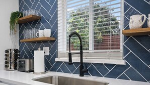 Une cuisine avec un évier en acier inoxydable, un robinet noir et une fenêtre avec des stores. On y trouve des étagères en bois où sont disposés des verres et une plante.