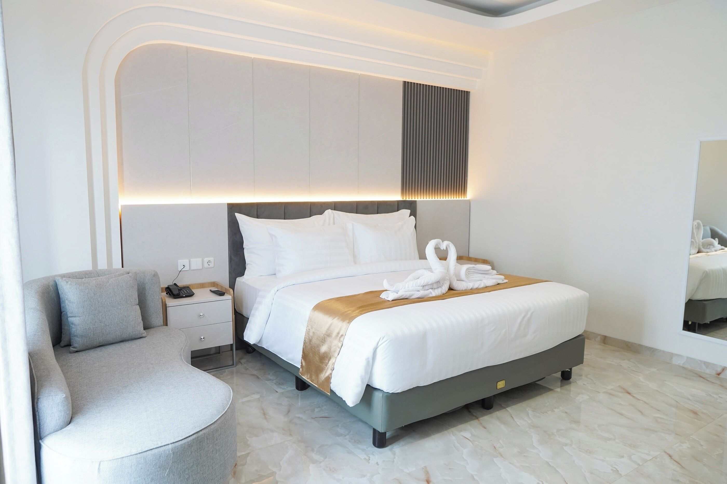 A modern hotel room with a curved gray sofa, a large bed with white linens and a golden throw, and a nightstand with a phone and charger.