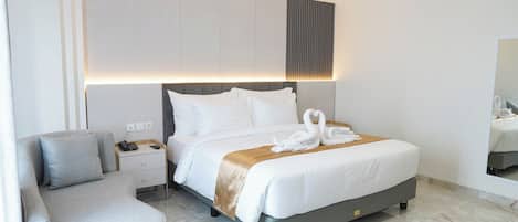 A modern hotel room with a curved gray sofa, a large bed with white linens and a golden throw, and a nightstand with a phone and charger.