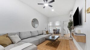 Un salon moderne avec un canapé d’angle gris, une table basse, un miroir rond et un téléviseur à écran plat fixé au mur.