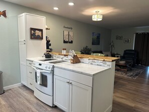 A compact kitchen with a built-in oven, a stovetop, and a small island with a sink. There are wall-mounted shelves and a painting above the island. - Family friendly townhouse style unit with a private deck and backyard!   (Anchorage)