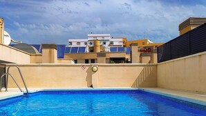 Une piscine avec un panneau indiquant qu’il est interdit de plonger, entourée d’immeubles dotés de balcons, sous un ciel dégagé.