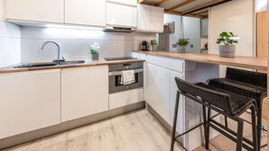 Une cuisine moderne avec des meubles blancs, un évier en acier inoxydable et un four encastré. On trouve deux plantes en pot sur le plan de travail et une chaise en osier noir.