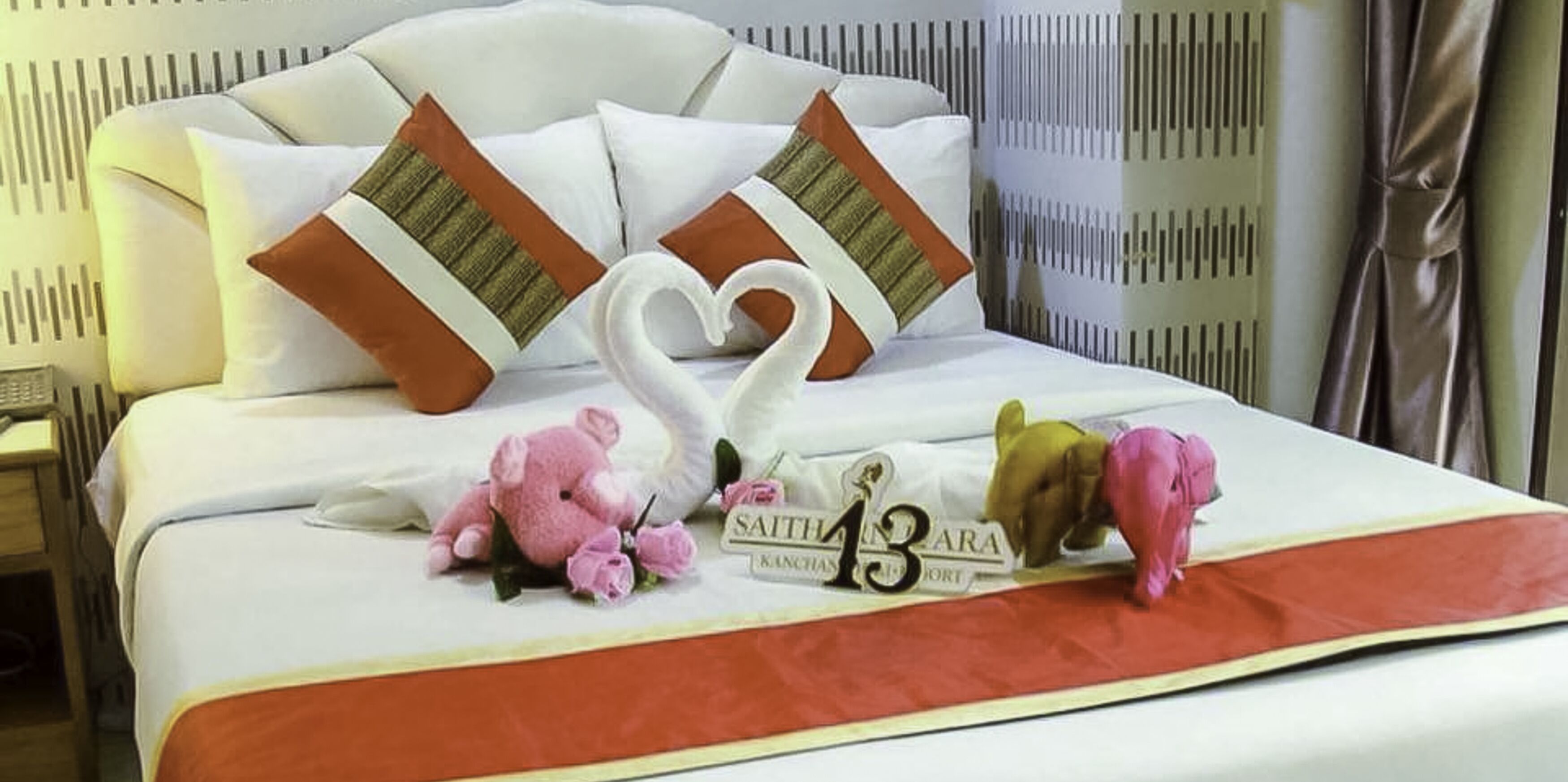 A neatly made bed with a decorative swan pillow arrangement, a number '13' sign, and a 'SAITHARA' sign on the bedspread.
