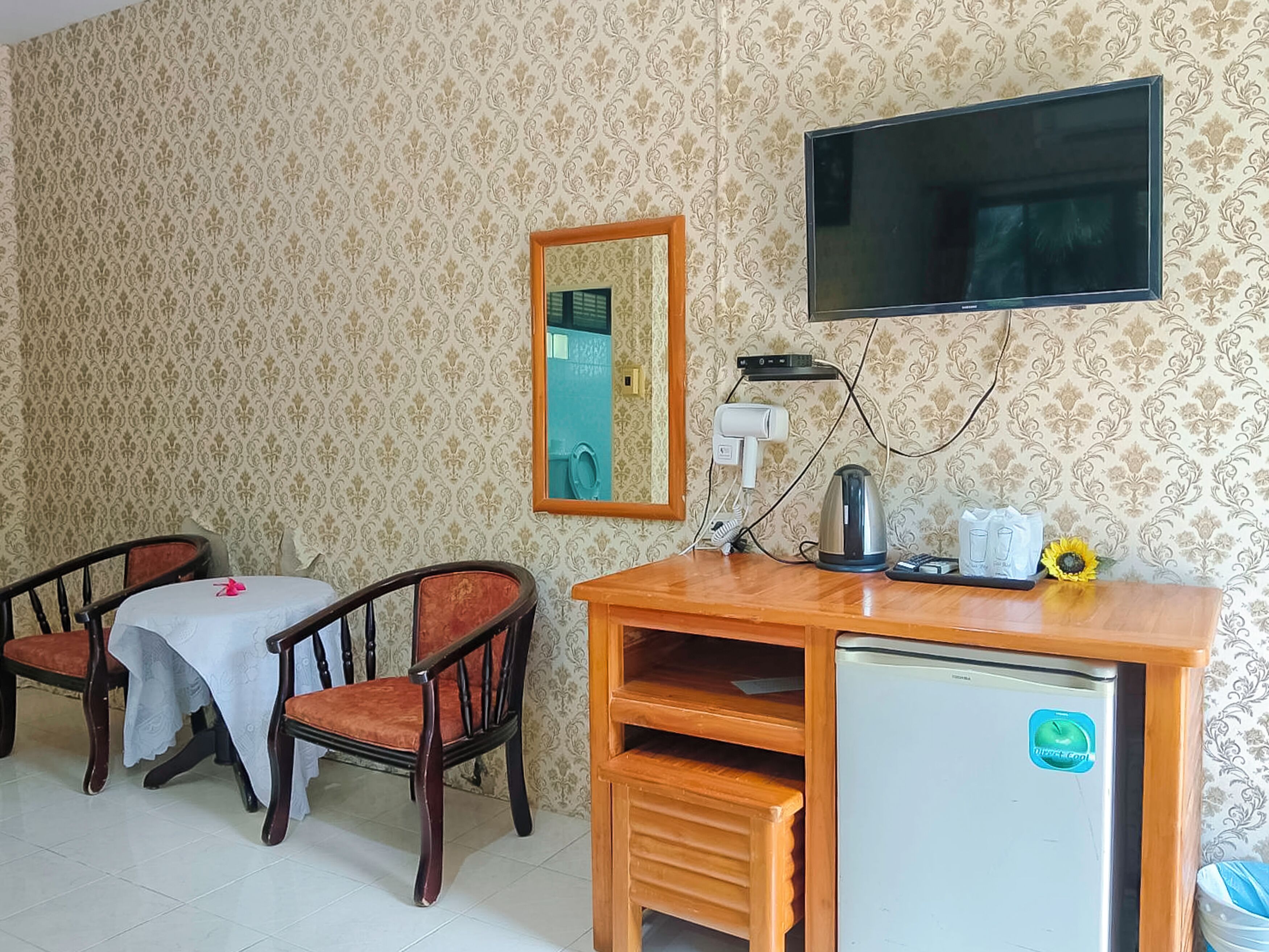 Een kamer met een televisie, een kleine koelkast, een tafel met stoelen en een spiegel aan de muur.