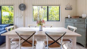 Un coin repas avec une table en bois et des chaises blanches, un vase rempli de fleurs, une horloge et une vue sur les arbres à travers de grandes fenêtres.