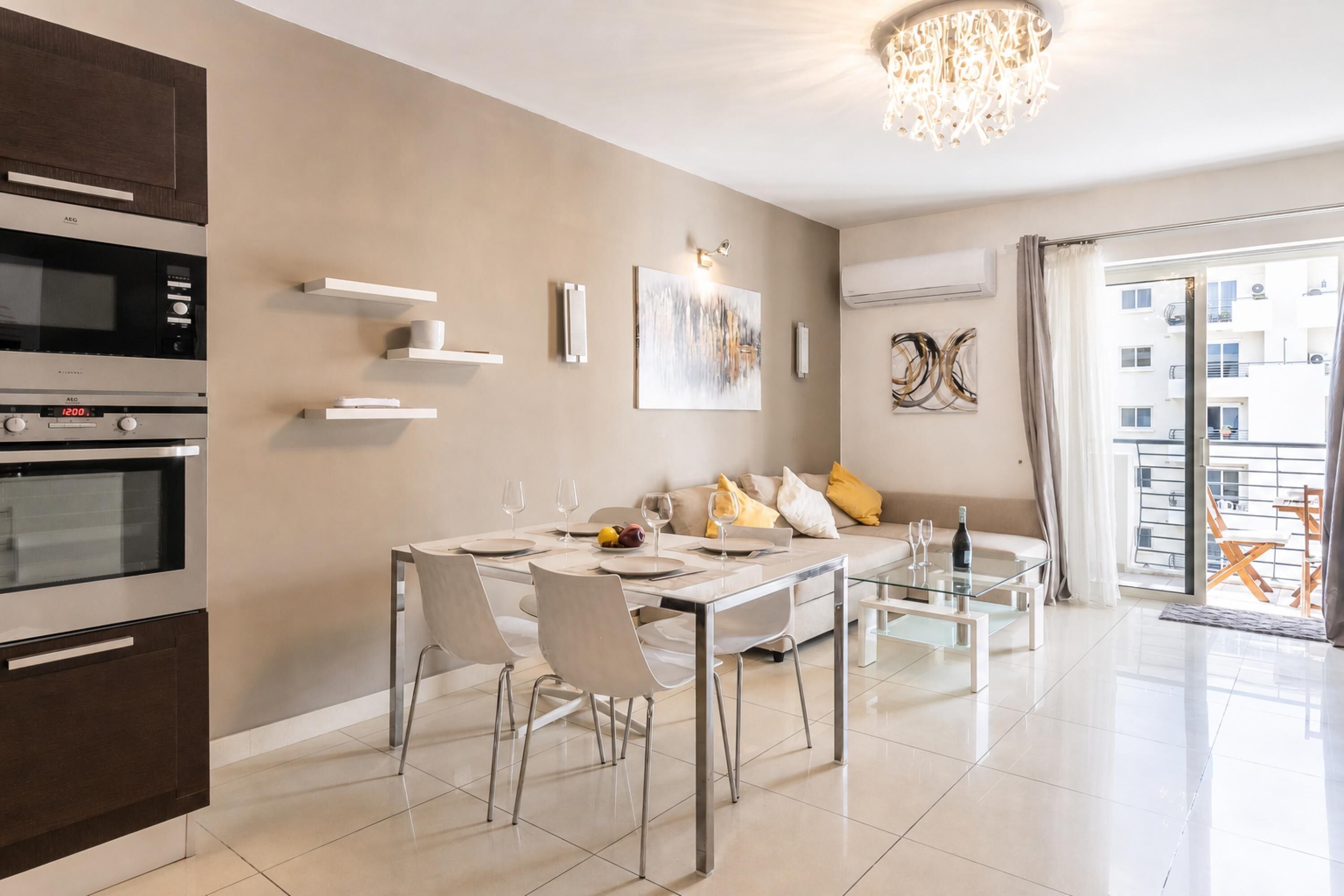 A modern kitchen with built-in appliances, a dining area with a glass table and chairs, and a living space with a sofa and artwork.