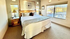 Een slaapkamer met een groot bed, een plafondventilator, een nachtkastje, een lamp en een raam met uitzicht op een gebouw en bergen.