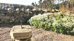 Eine Steinsäule mit Sonnenuhr in einem Garten mit weißen Blüten und Bäumen im Hintergrund.
