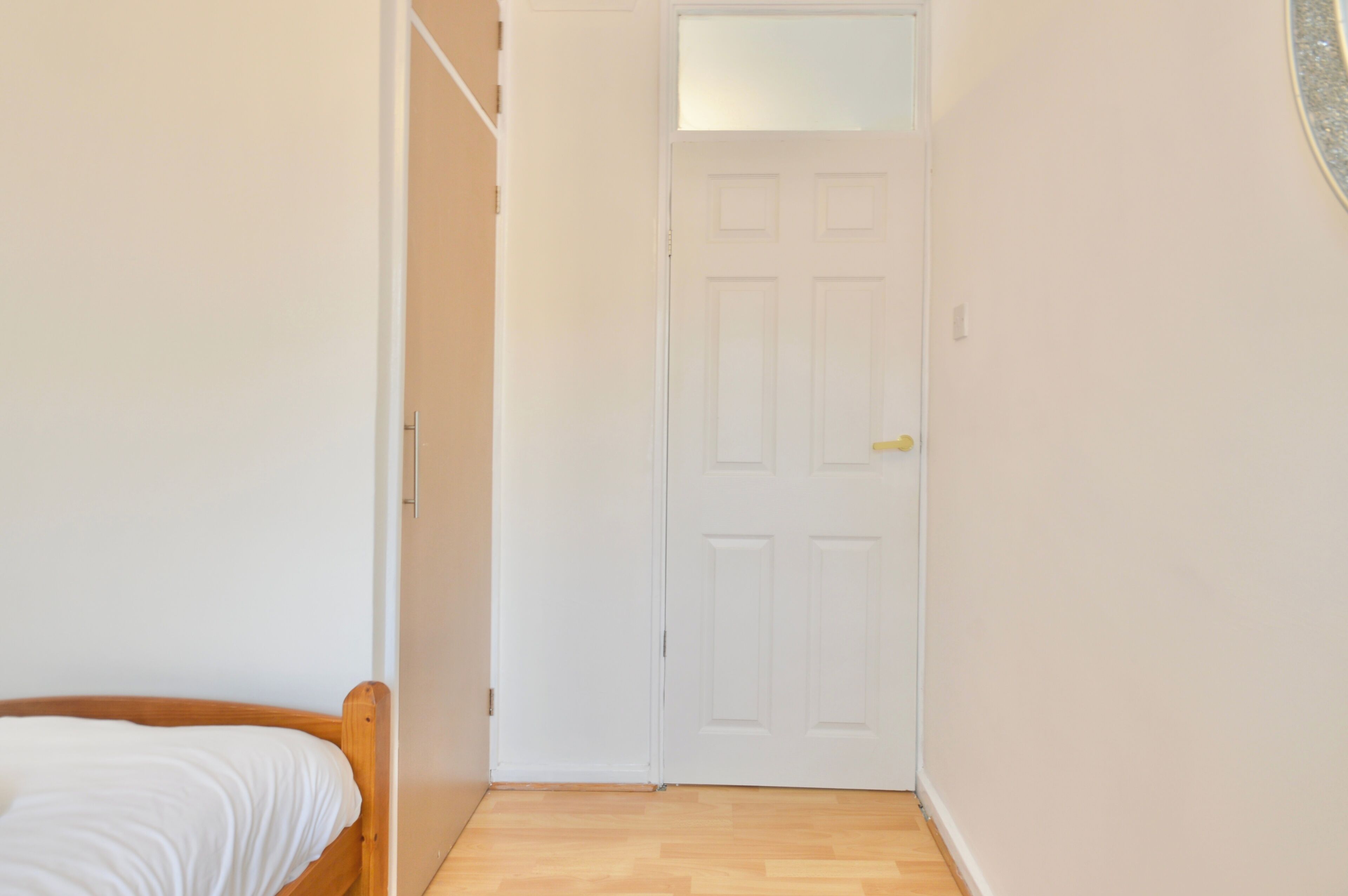 A narrow hallway with a wooden floor, a white door with a small mirror, and a bed with a white sheet.