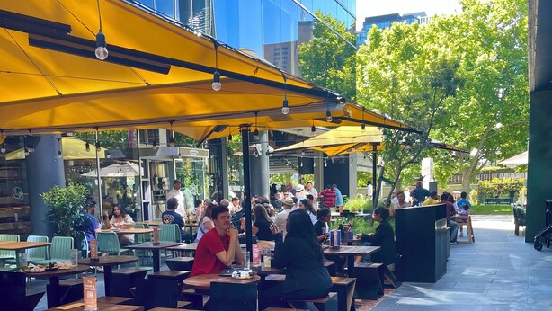 An outdoor dining area with wooden tables and chairs, covered by yellow umbrellas, and a modern building in the background. - Melbourne Riverside Retreat with Pool. Central Location. Family-Friendly. (Southbank)