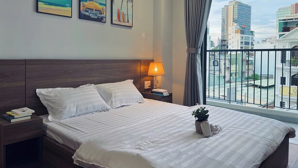 A hotel room with a large bed, a nightstand with a lamp, and a window with a view of the city. - SenseHome - 5 mins to Bui Vien street  (Ho Chi Minh City)