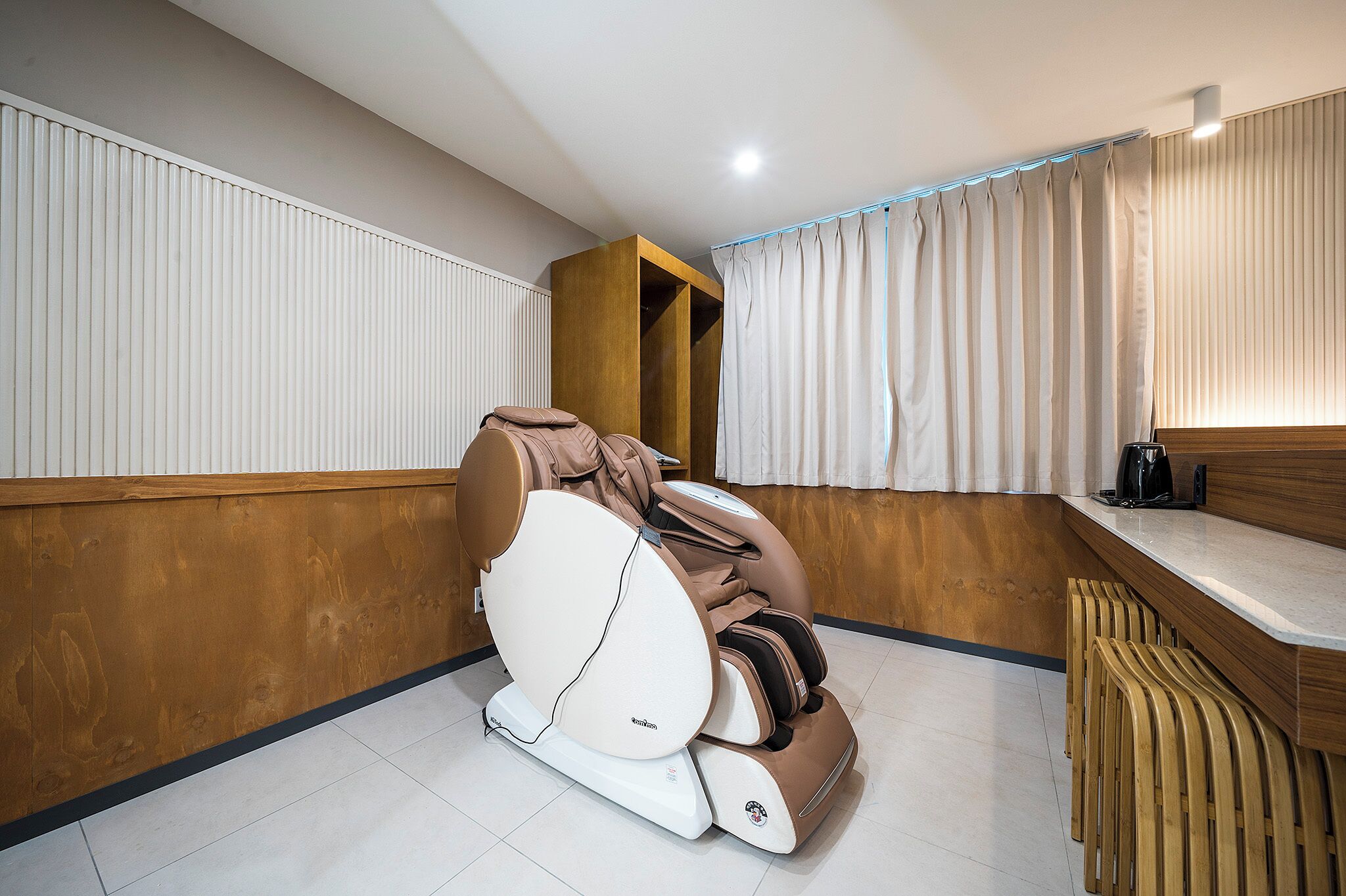 Ein Behandlungsraum für Massagen mit Massagegerät, Holzvertäfelung, einer Theke mit Kaffeemaschine und einem verhängten Fenster.