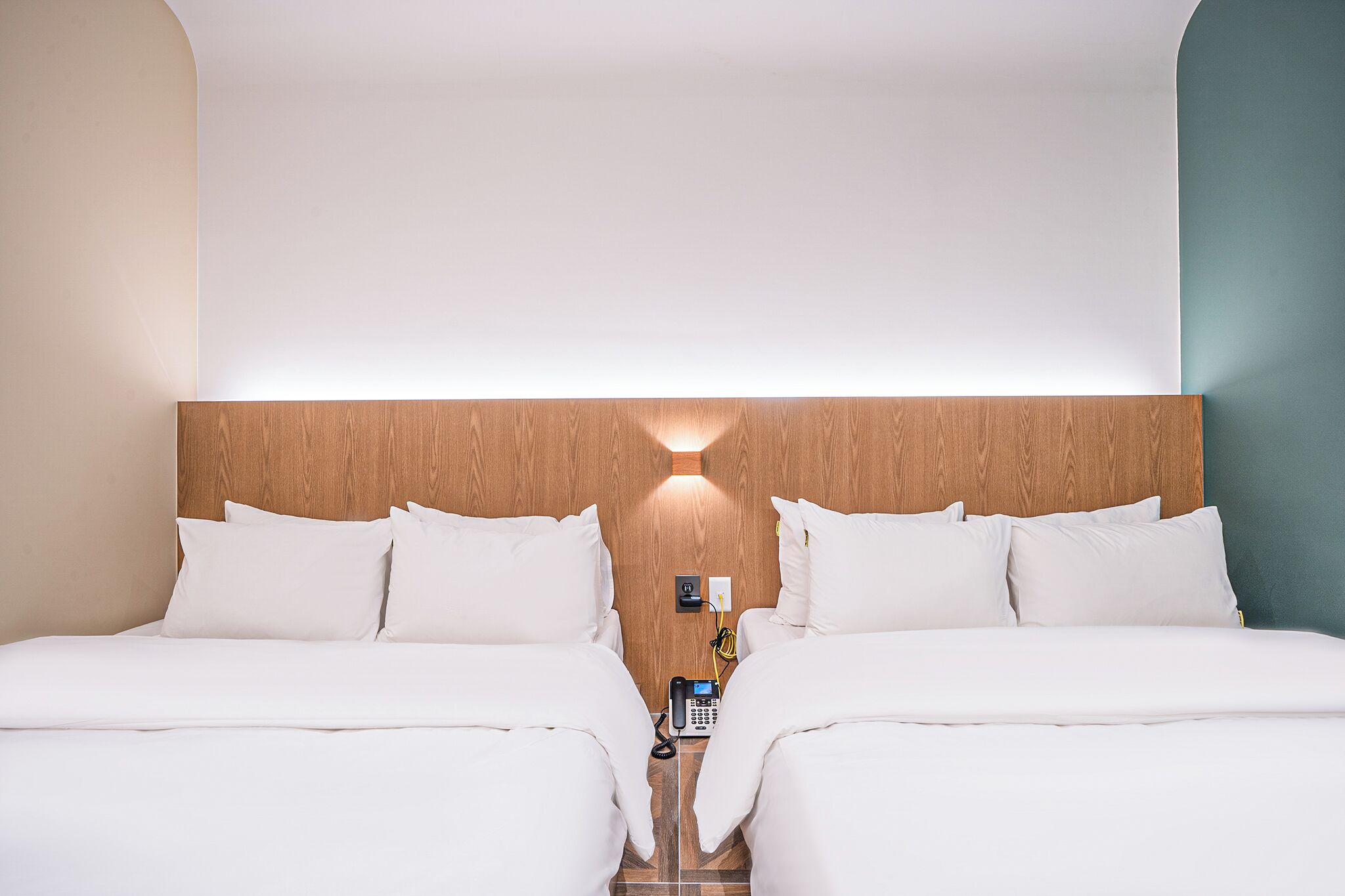 Habitación de hotel con cabecera de madera, dos camas con ropa blanca, una mesita de noche con teléfono y una lámpara fijada a la pared.