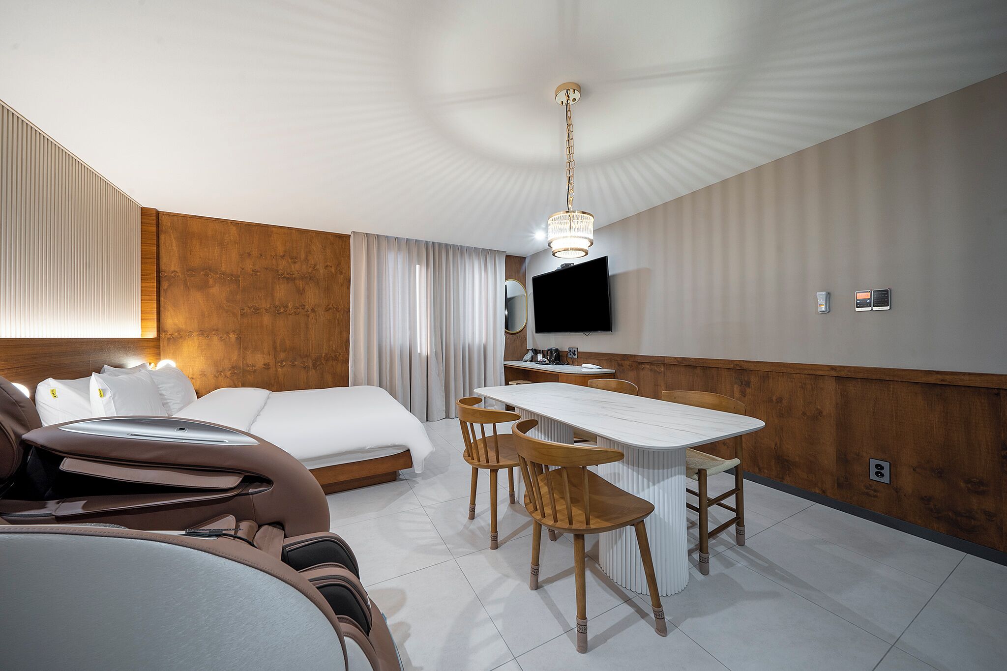 Una habitación de hotel moderna con una cama, una mesa de comedor con sillas, un televisor y una lámpara en el techo.