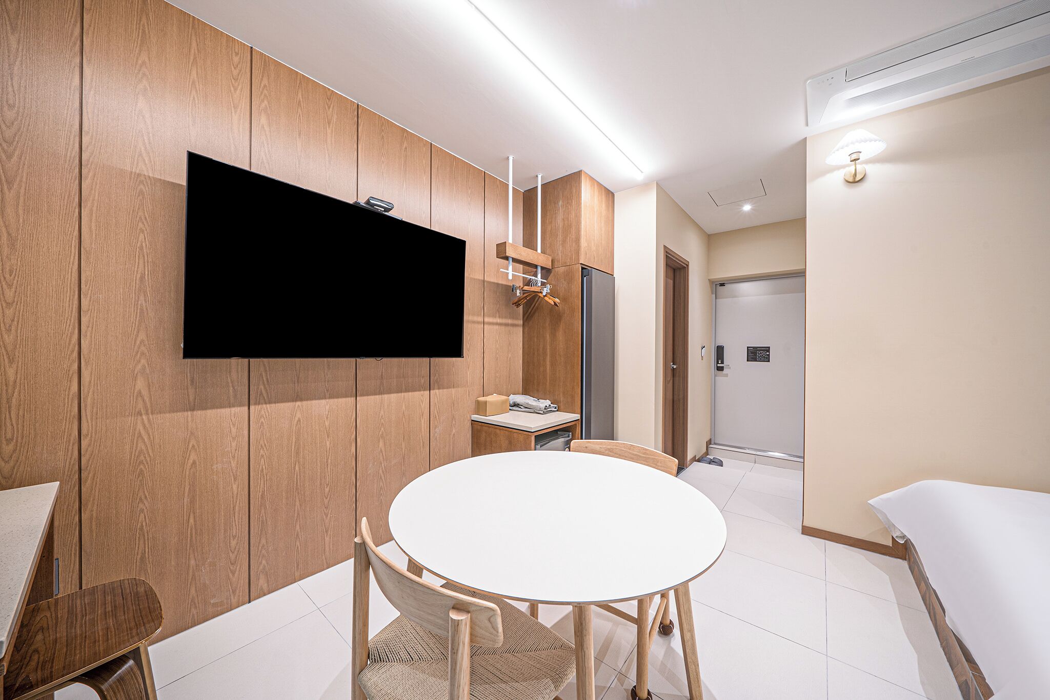 A modern hotel room with a round dining table, wooden panel walls, a flat-screen TV mounted on the wall, a small kitchen area, and a separate sleeping area.