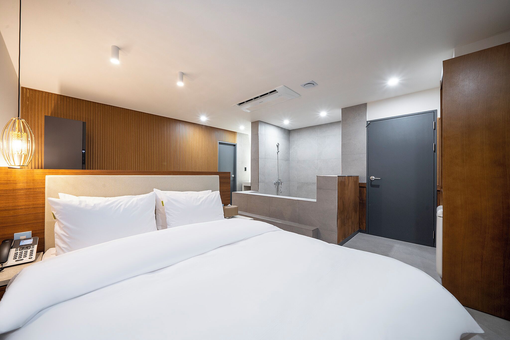 Una habitación de hotel moderna con una cama grande, teléfono en la mesita de noche, pared de paneles de madera, televisor integrado, zona de ducha y una puerta oscura.