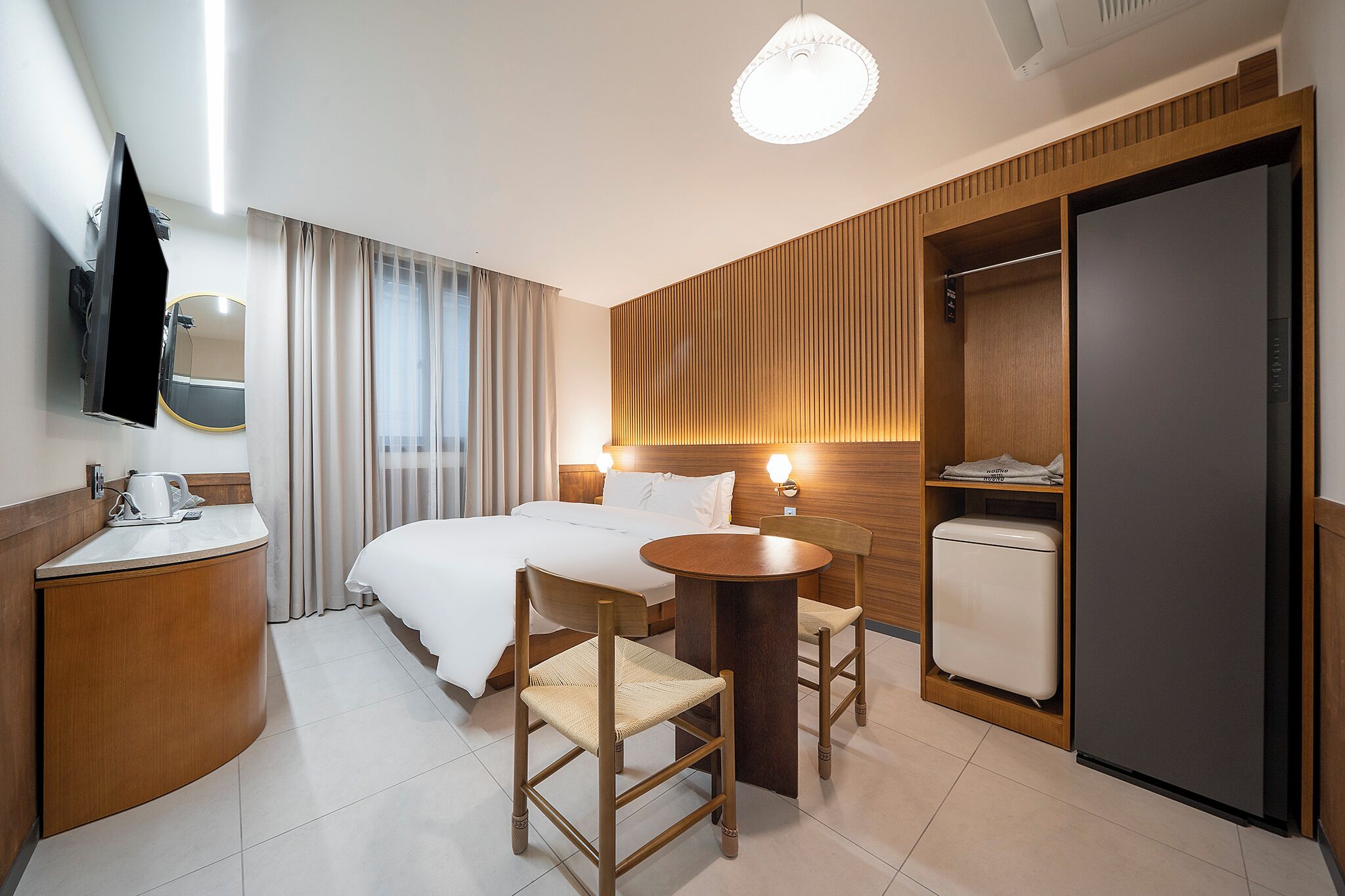 A hotel room with a bed, bedside table, chairs, a small table, a mirror, a wardrobe, and a television.