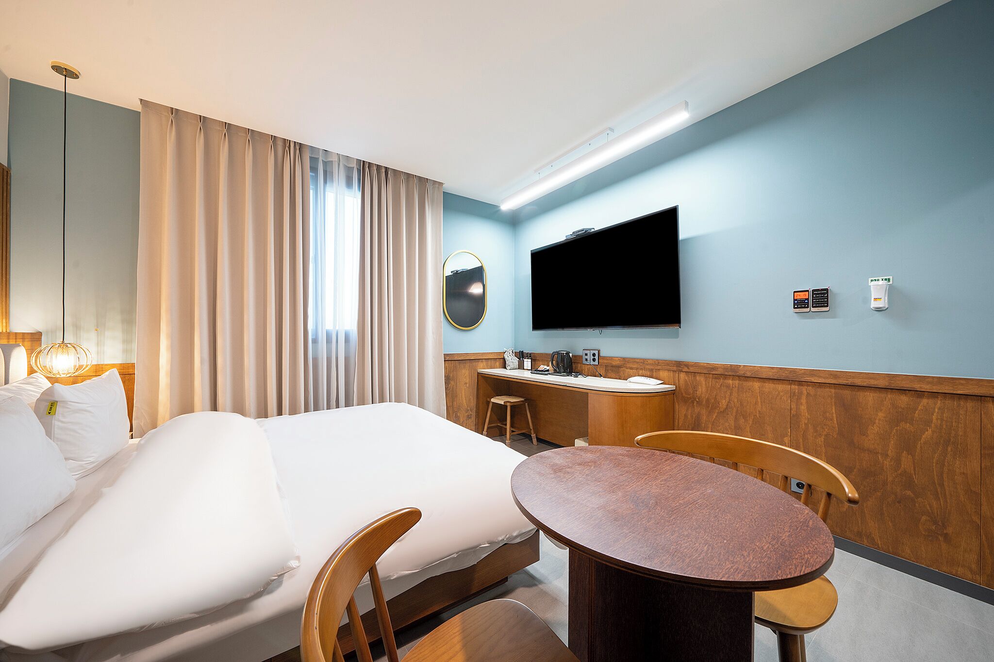 A hotel room with a bed, a desk, a chair, a round table, a TV mounted on the wall, and a window with curtains.