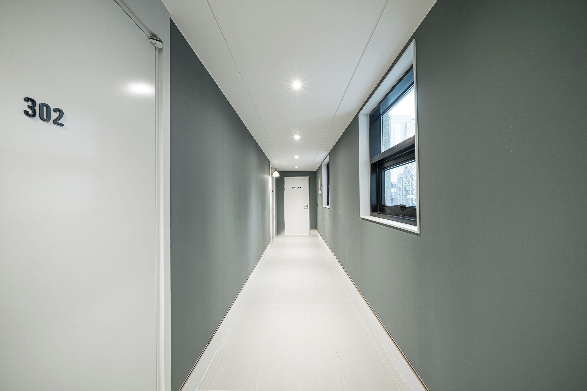 A narrow, well-lit hallway with a number "302" on the left wall, a window on the right, and a door at the end.