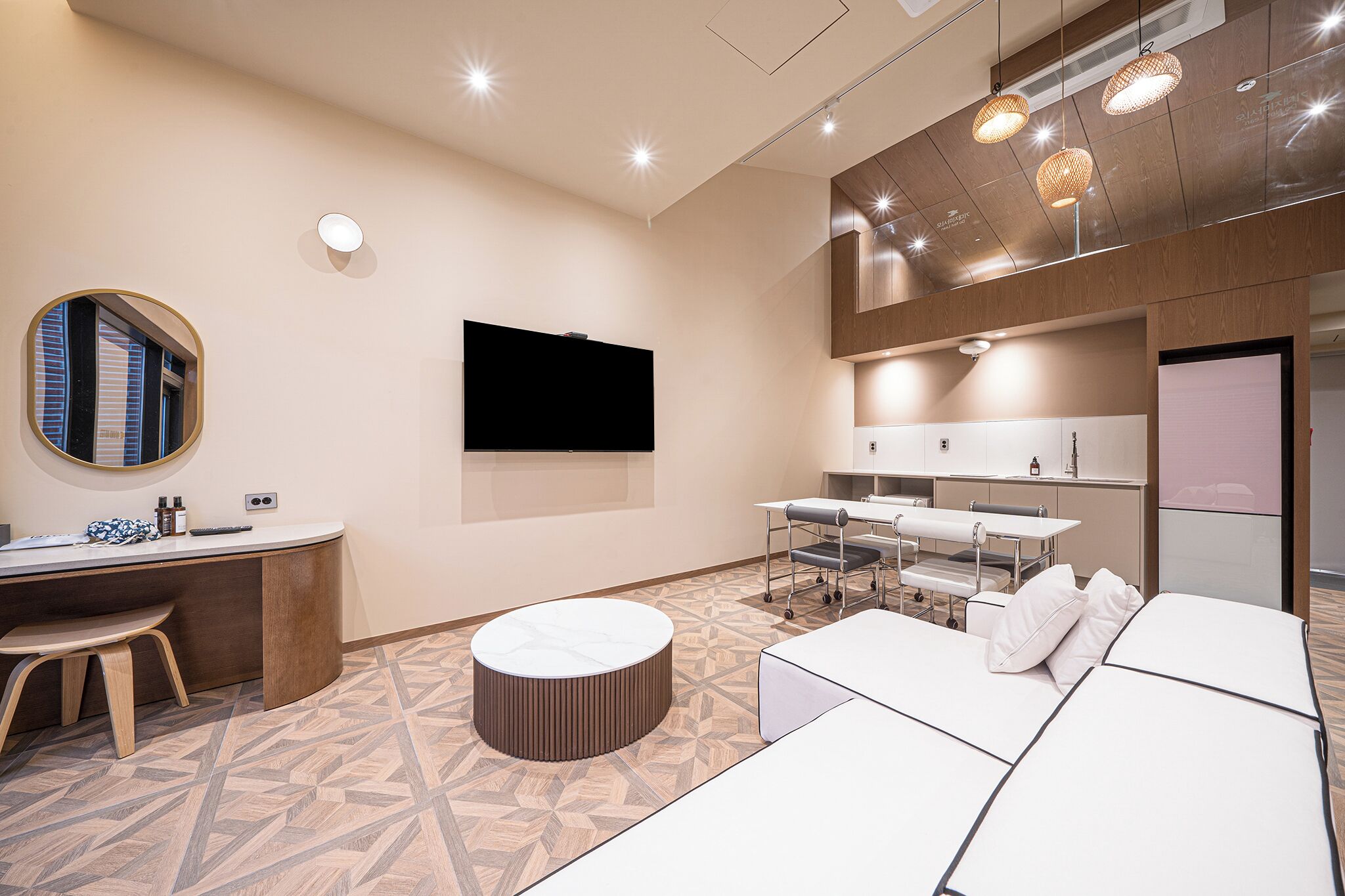 A modern hotel room with a flat-screen TV, a desk with a chair, a round ottoman, and a sofa with pillows.