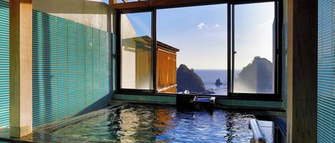 Un onsen japonais traditionnel doté d’une grande fenêtre offrant une vue sur la mer et les falaises rocheuses.