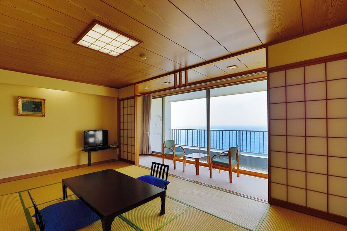 Une pièce de style japonais traditionnel, avec un sol recouvert de tatamis, une table basse et un téléviseur. Un balcon avec vue sur la mer est visible à travers de grandes portes coulissantes.