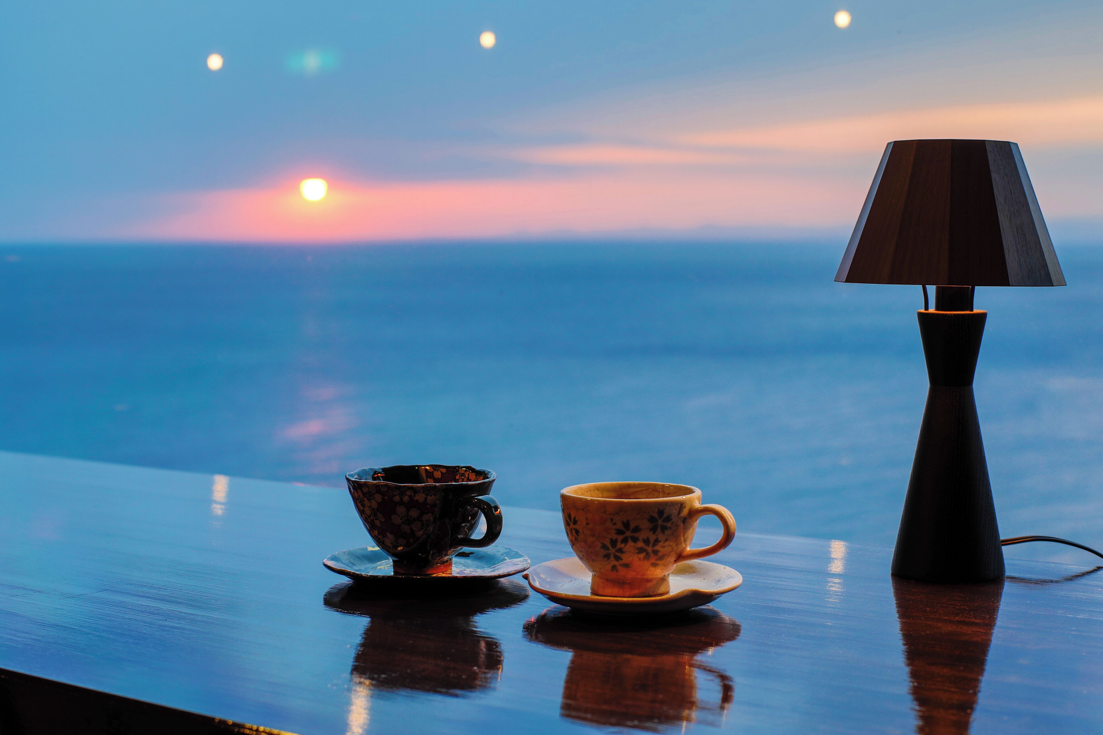 Una mesa con dos tazas y una lámpara, con vista a la puesta de sol sobre el océano.