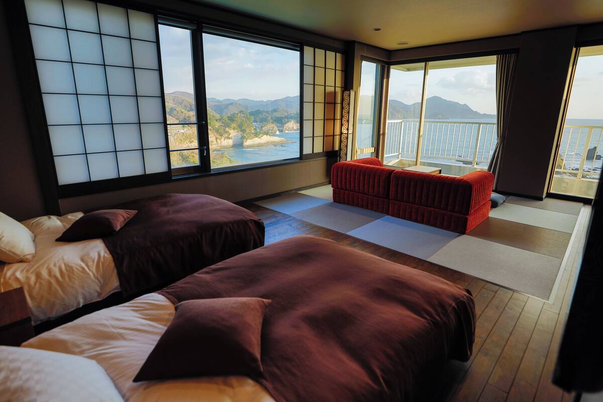 Une chambre d’hôtel avec deux lits, un canapé rouge, une grande fenêtre donnant sur la mer et les montagnes, et une porte coulissante donnant sur un balcon.
