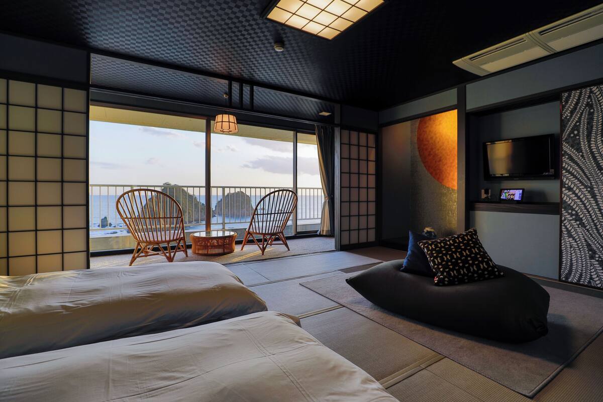 Une chambre d’hôtel moderne dotée d’une grande fenêtre, de deux fauteuils en bois, d’un canapé et d’un téléviseur à écran plat.
