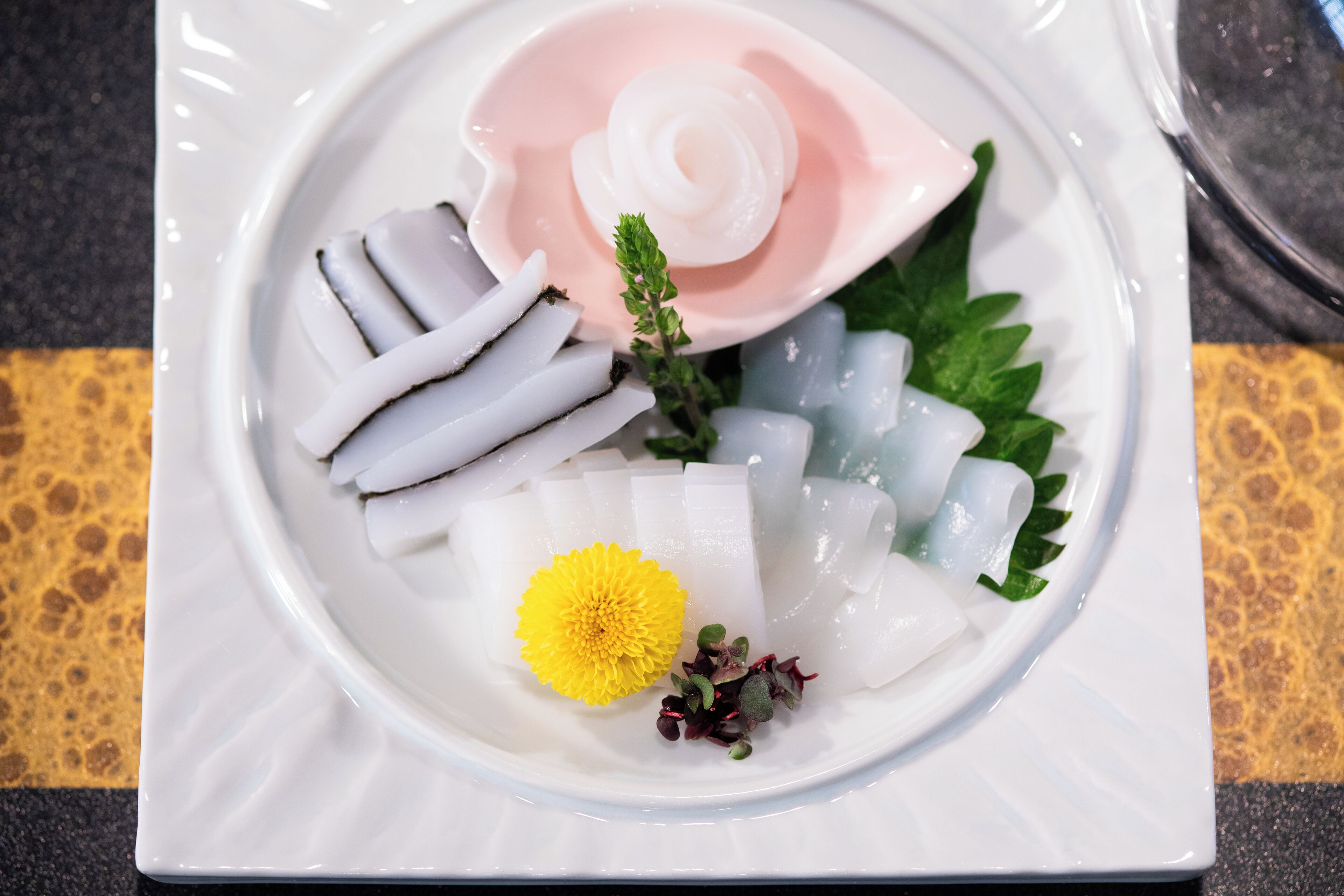A white plate with sliced white fish, a pink flower, and green leaves.