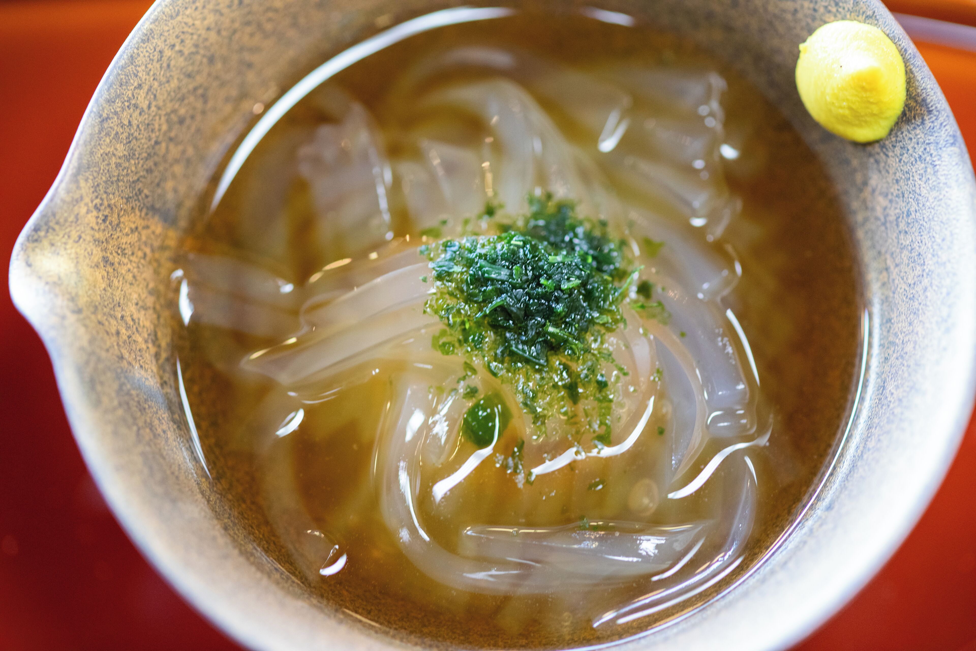 Un bol de soupe claire à base de bouillon, de nouilles et d’herbes fraîches, servi dans un bol en céramique avec une tranche de citron à côté.