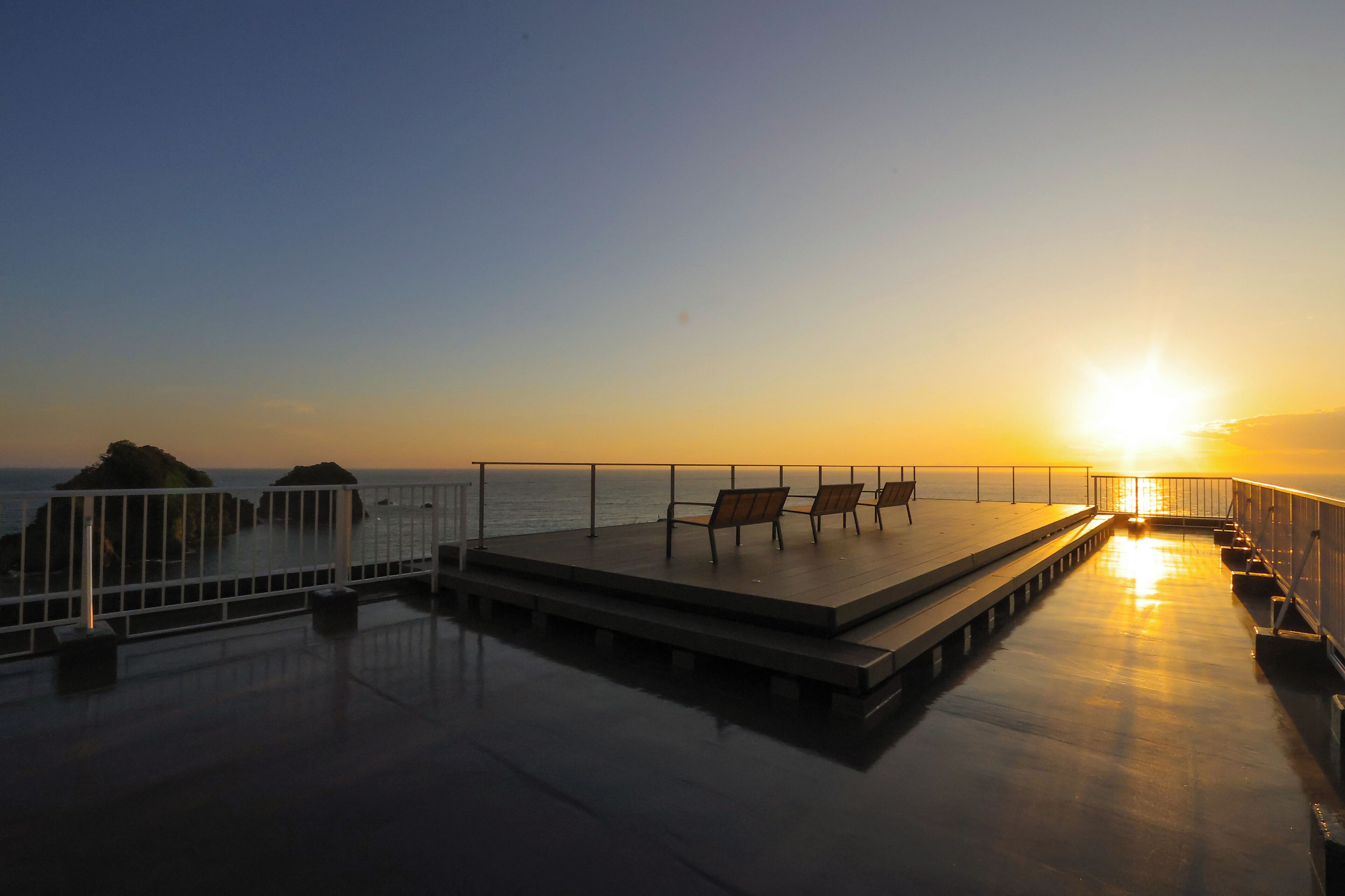 A sunset view from a deck with benches and a railing overlooking the sea.