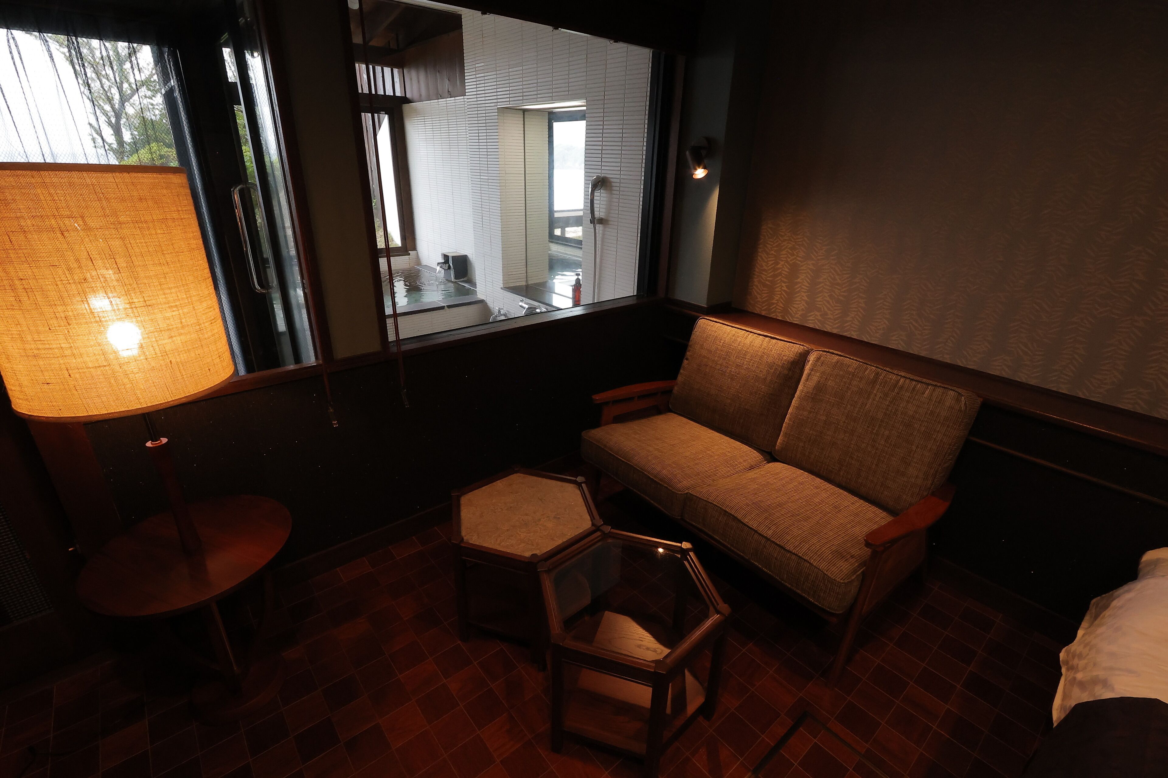 Una habitación tenuemente iluminada con un sofá, una mesita y una silla hexagonal. Una ventana muestra un baño con un espejo y una ducha.