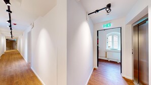 Un couloir moderne aux murs blancs, au plancher en bois et doté d’un éclairage fixé au plafond. Un panneau indiquant la sortie de secours est visible sur le mur de droite.