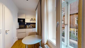 Une cuisine compacte dotée d’appareils électroménagers modernes, une table à manger et une vue sur un bâtiment en briques, visible à travers une fenêtre.
