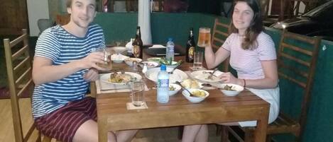Two people seated at a wooden dining table with plates of food, glasses, and bottles.