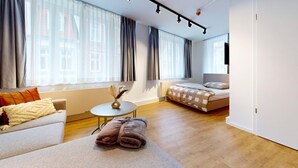A modern hotel room with a sofa, a small round table, a bed with a checkered duvet, and large windows with curtains.
