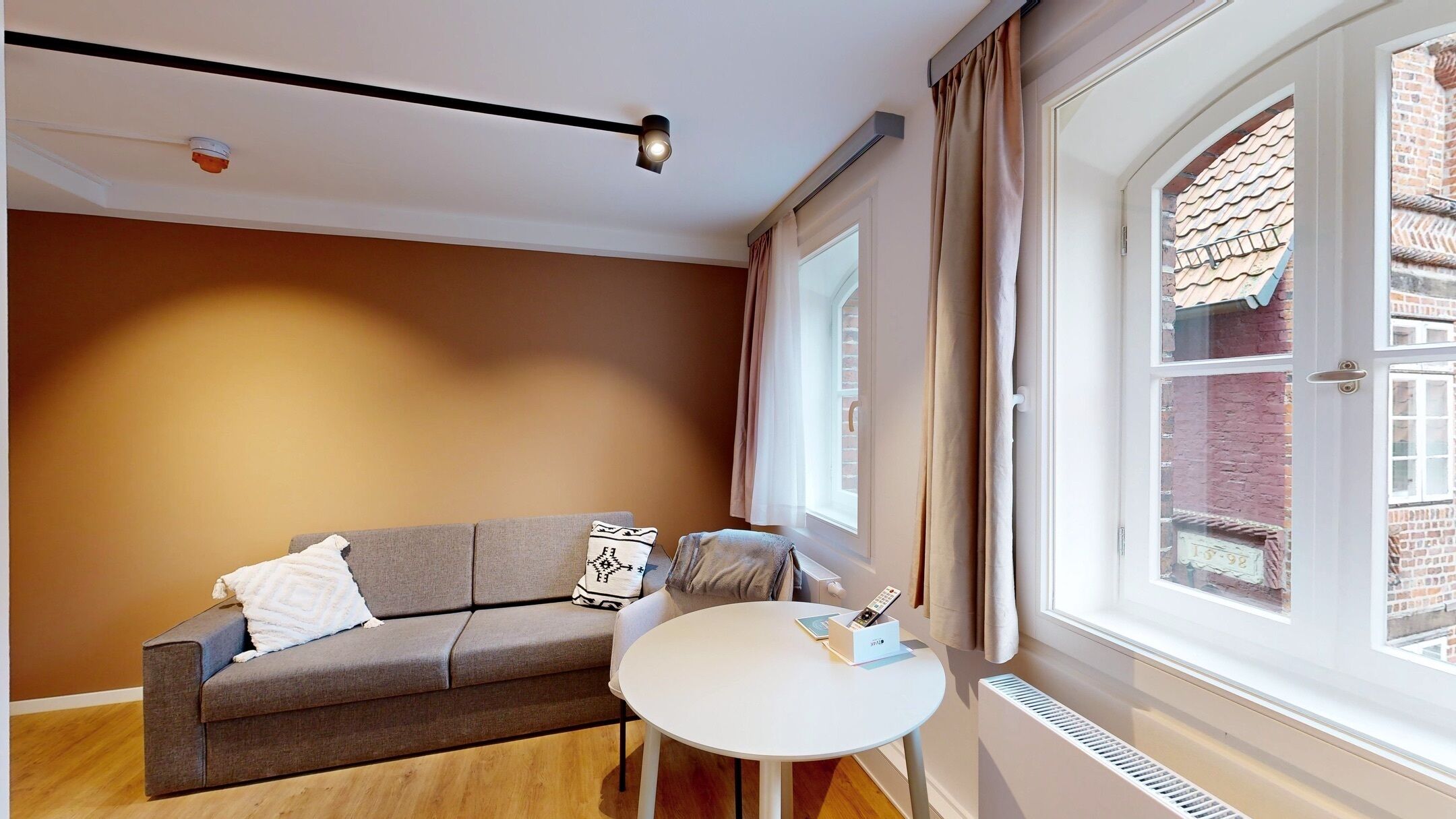 Ein modernes Wohnzimmer mit grauem Sofa, einem kleinen weißen Tisch und einem Fenster mit Vorhängen.