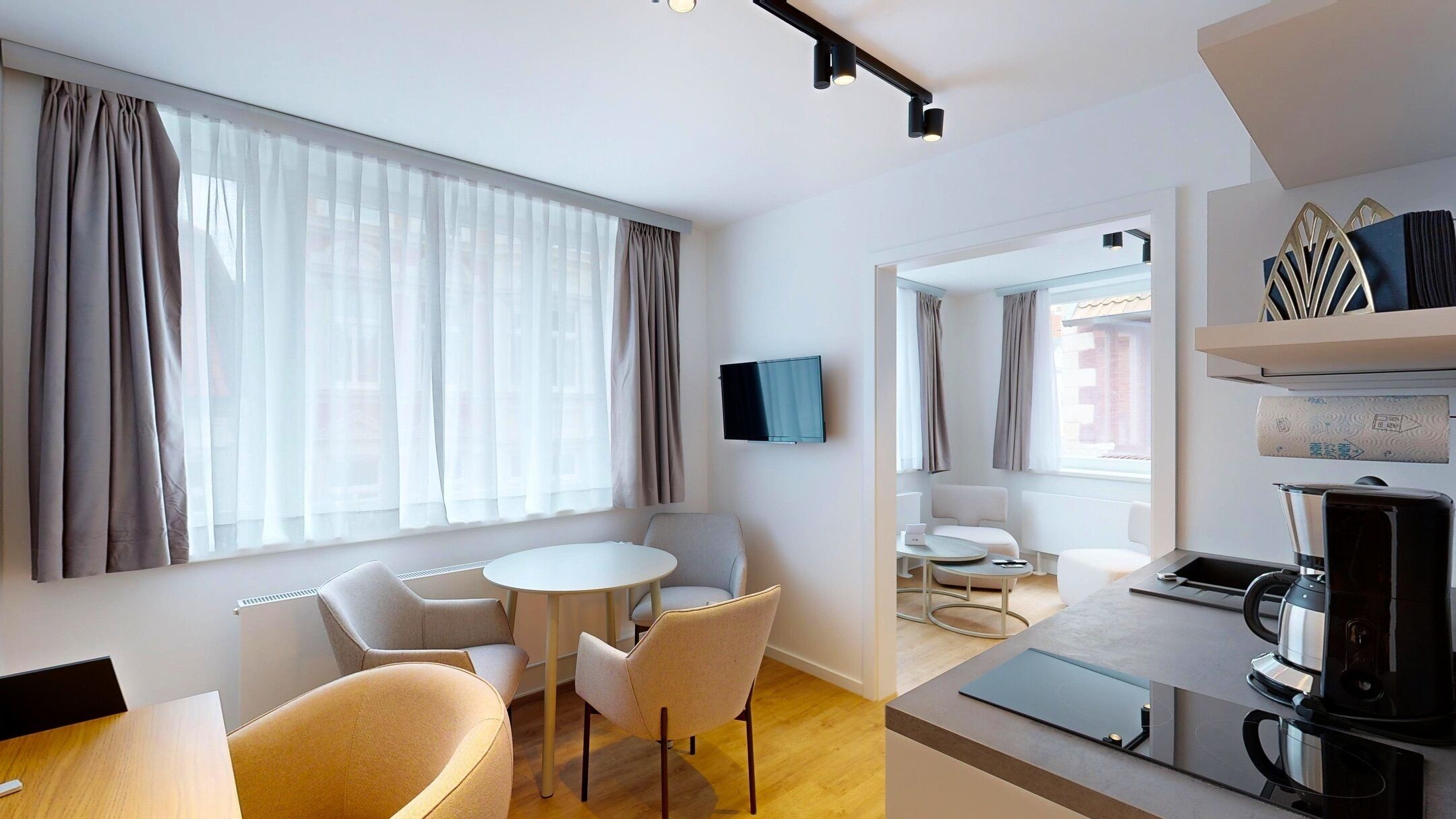 A modern hotel room with a dining area, a TV mounted on the wall, and a kitchen area with a coffee maker.