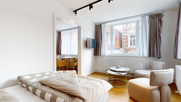 A modern hotel room with a bed, a small round table, a sofa, and a TV mounted on the wall. - Apartment 9 | Boardinghouse Lüneburg No8A (Lüneburg)